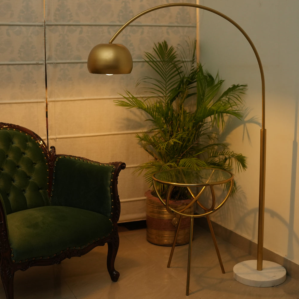 a lifestyle image of an arched tall metallic floor lamp with a marble base, placed in a living room set-up.