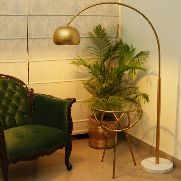 a lifestyle image of an arched tall metallic floor lamp with a marble base, placed in a living room set-up.