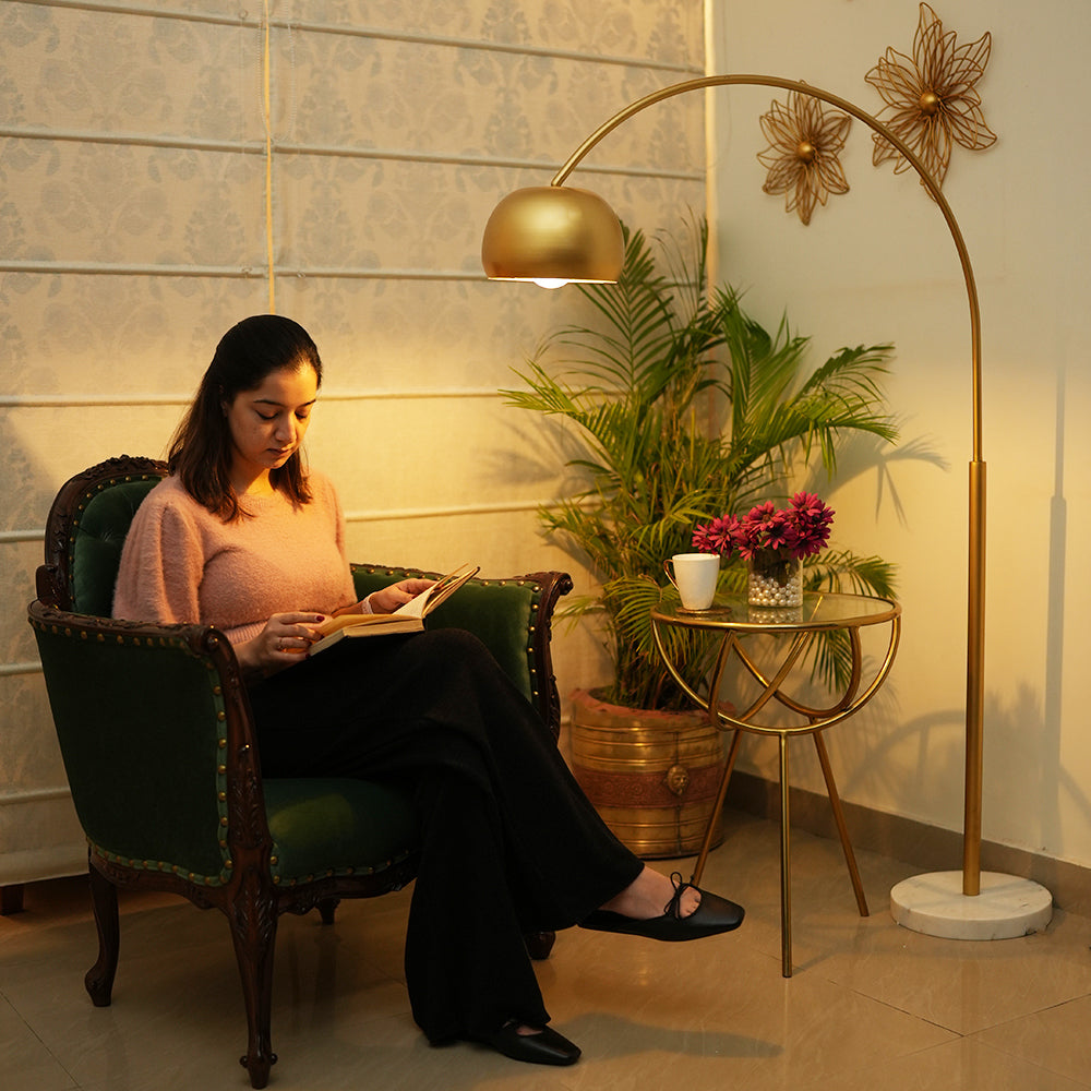 A lady sitting on an armchair and reading a book with her legs crossed, and a tall golden arched top floor lamp is lit next to her in a living room setup.