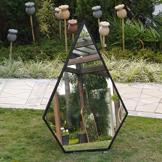 Front view of a tall metallic black pentagon framed mirror placed in a garden.