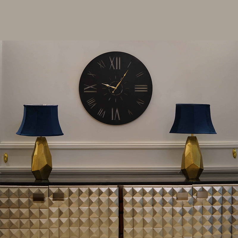 Large black wall clock with roman numerals with two golden lamps on either side of the clock, placed on a console.