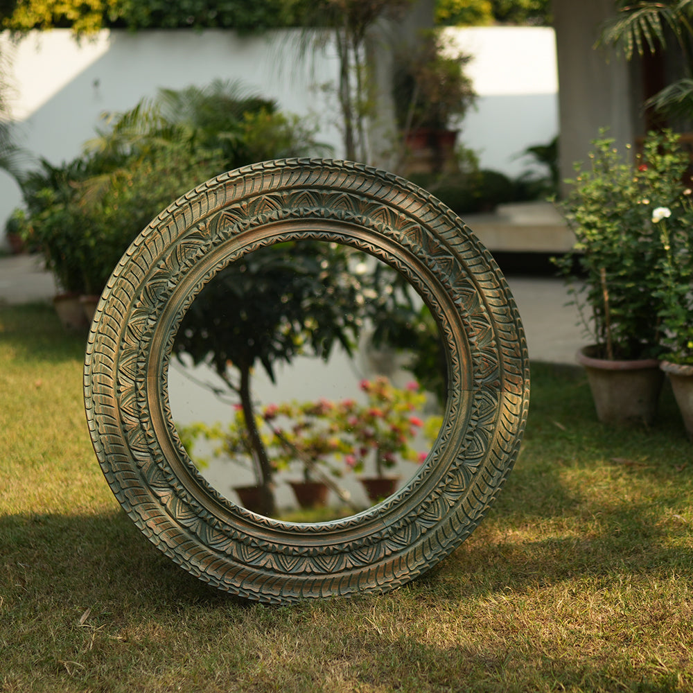 A large round mirror made from hand-carved mango wood with intricate detailing, placed in a garden setup.