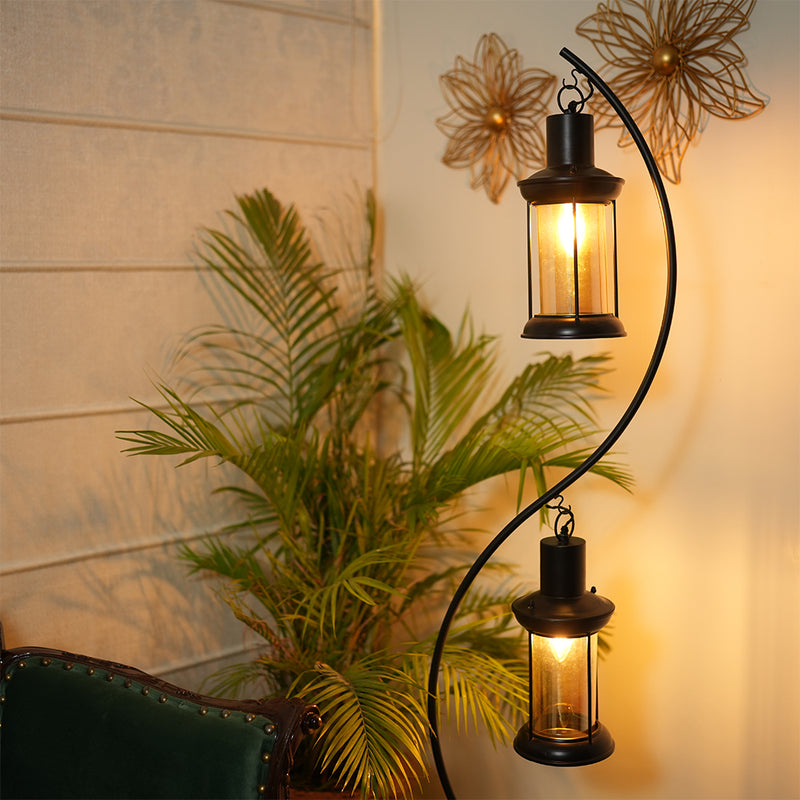 A floor lamp with two lanterns lit up in a living room setup, placed next to a green chair and plants on the background.