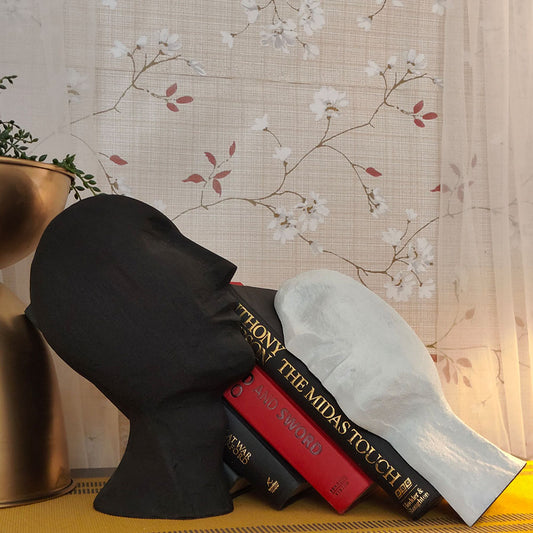 A bookend pair in the shape of an upward looking human face split in half resting on a stack of books.