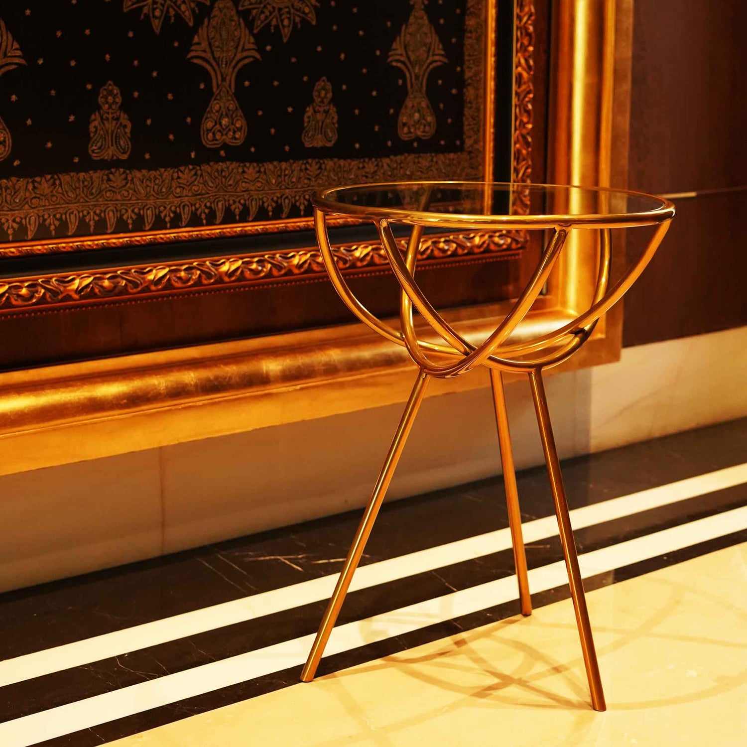 Close up view of a modern design golden colored metallic side table with a glass top in front of a large golden wall art.
