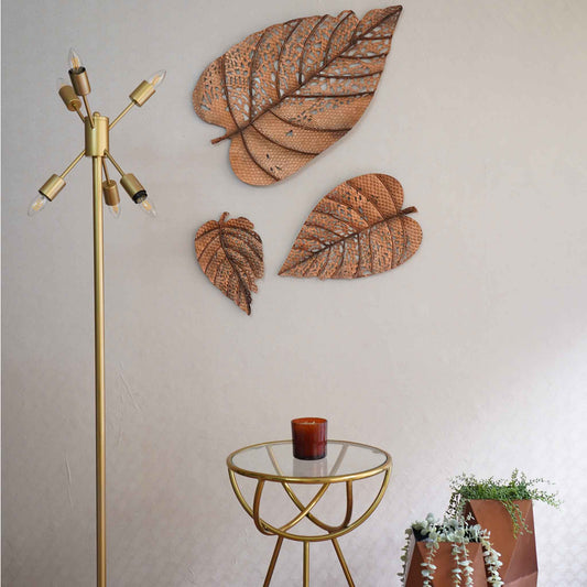 Set of 3 metallic leaves wall art displayed in a living room set up with a tall floor lamp, a table and two vases.