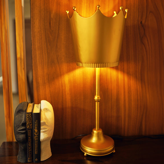 Imperial design golden metallic table lamp with a crown shaped shade, lit  up on a console table with a set of bookends placed next to it.