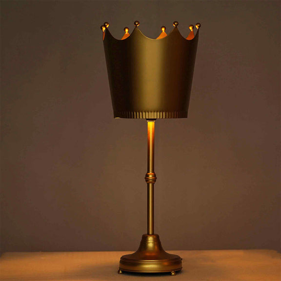 Imperial design golden metallic table lamp with a crown shaped shade lit up in ambient yellow light.