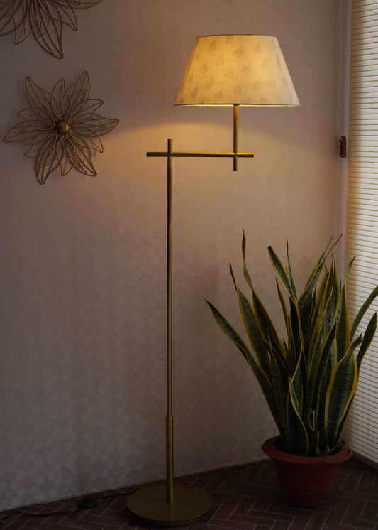 Tall, minimalistic design golden floor lamp with shade placed in front of a wall with wall art and next to a plant.