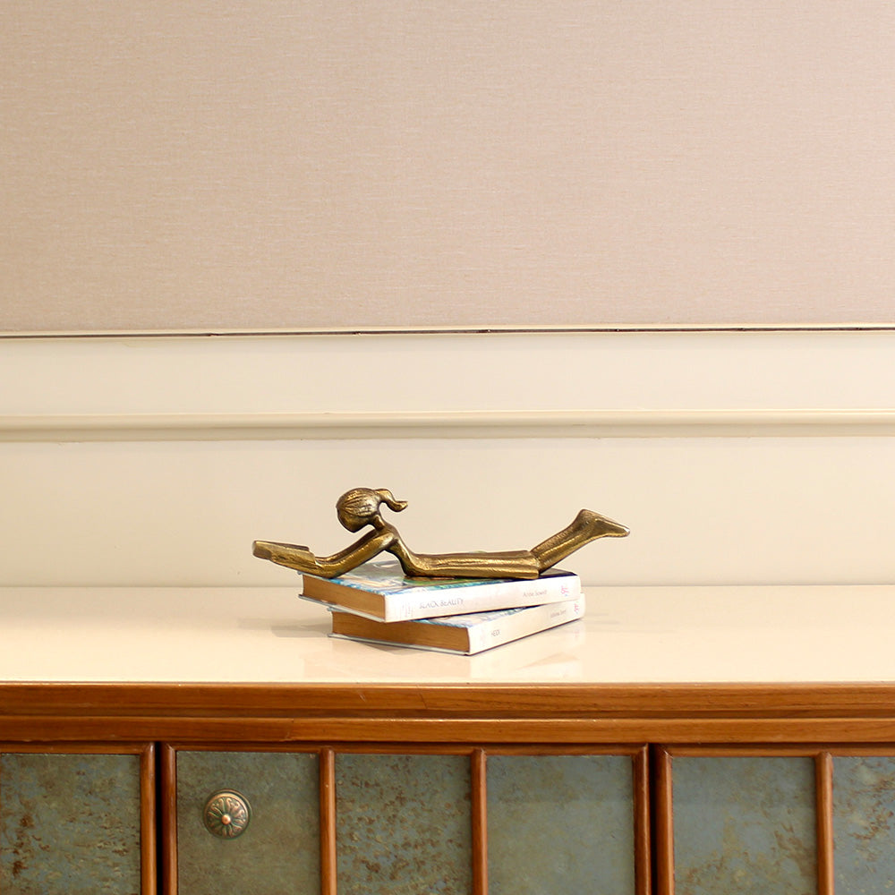 Table-top metallic decorative figurine with girl lying down and reading a book- side view.