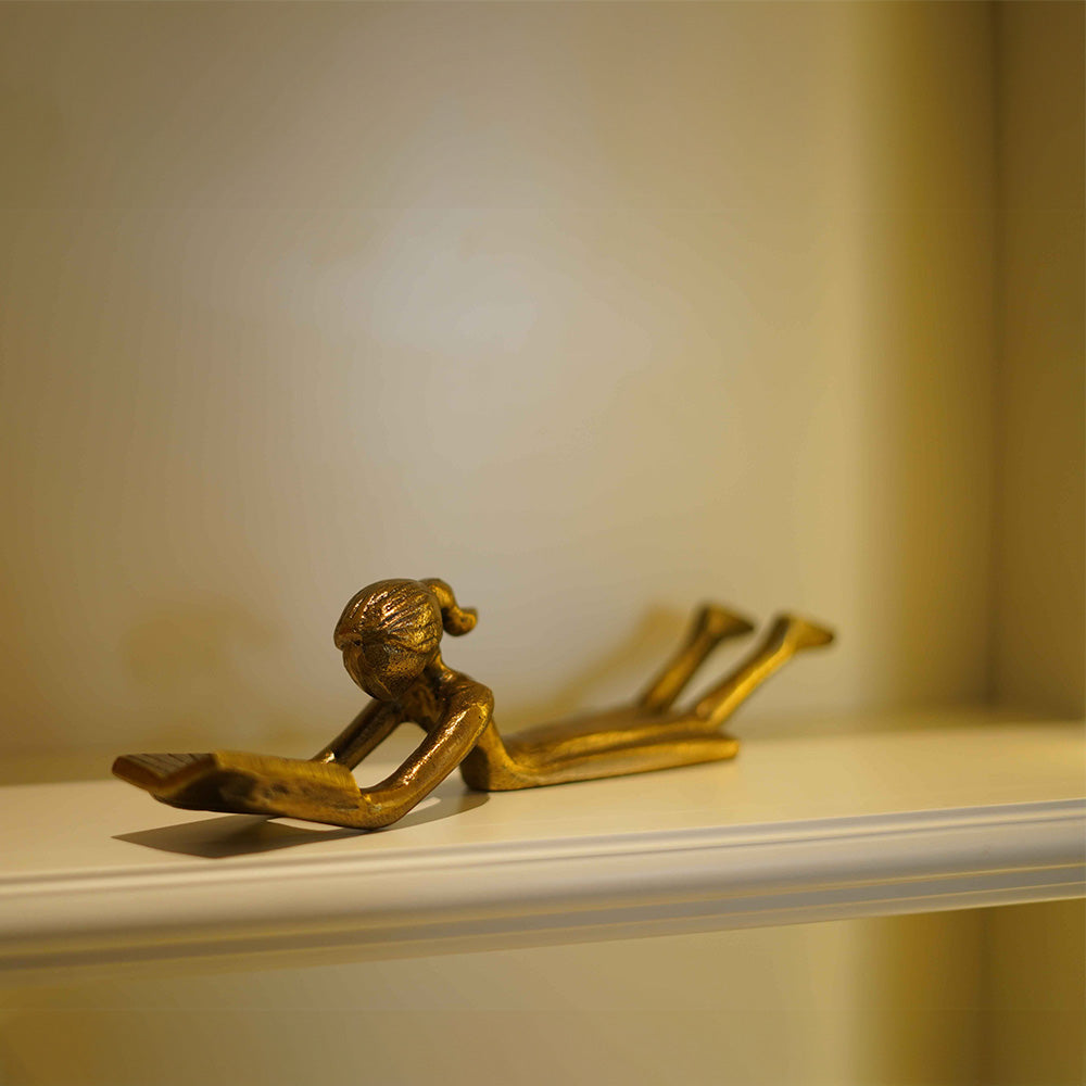 Table-top metallic decorative figurine of a girl lying down and reading a book- side view.