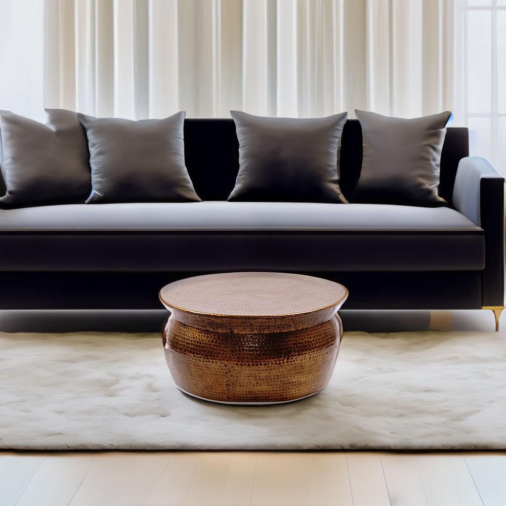 A metallic copper color round center table on the carpet of a living room with black sofa and white curtains in the background.