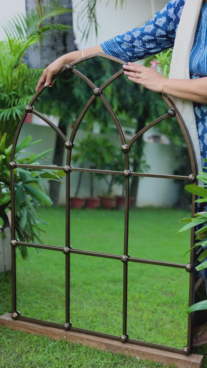 Video of a large metallic window shaped mirror placed in the garden and a female hand g=feeling the edges and curves of the mirror.