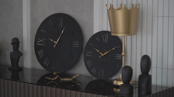 Zoomed in video of two large black wall clocks with roman numerals placed on a console table.