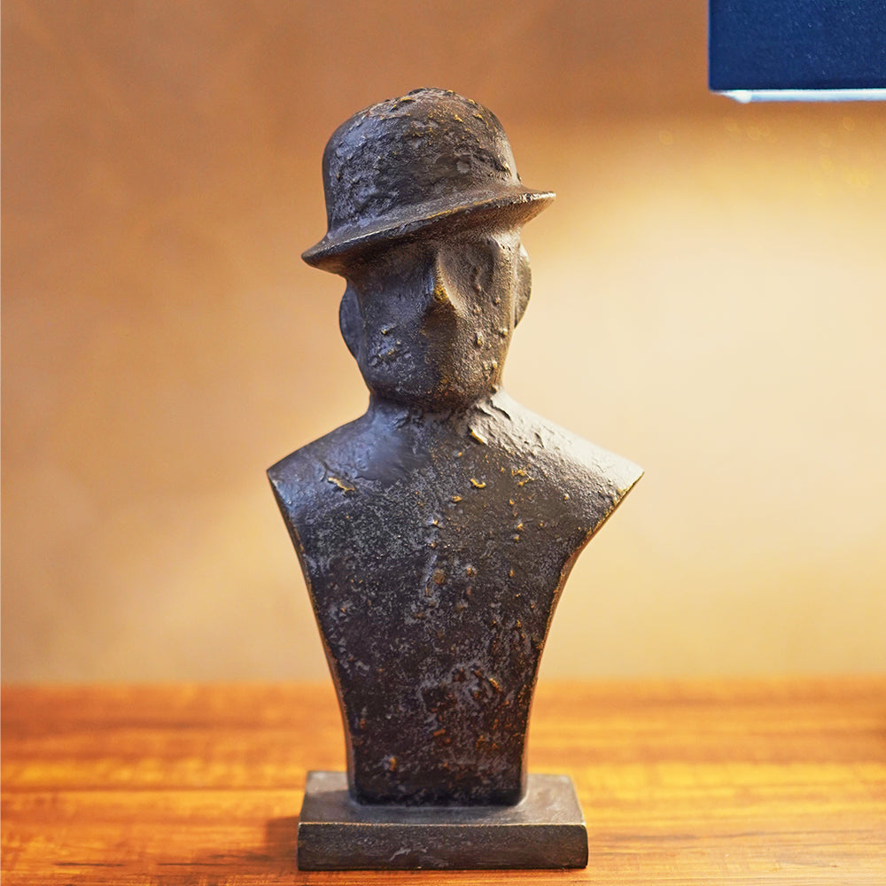 Front view of a black metallic table-top sculpture of a man wearing a tilted hat, placed on a side table. 