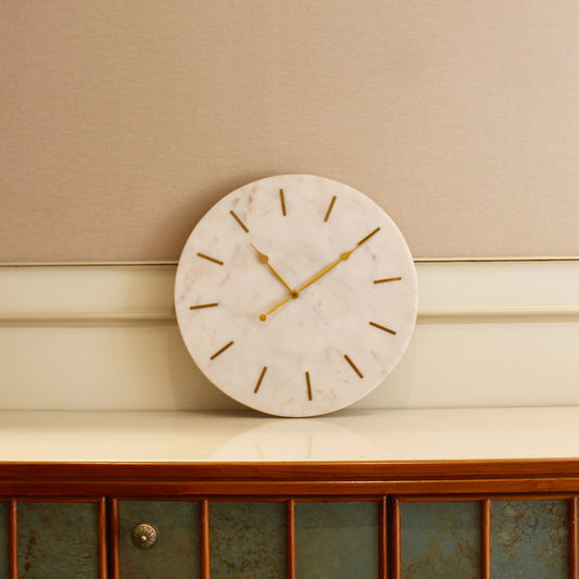 A large and heavy white marble clock with golden hands and hours marked with brass inlay placed on a console table.