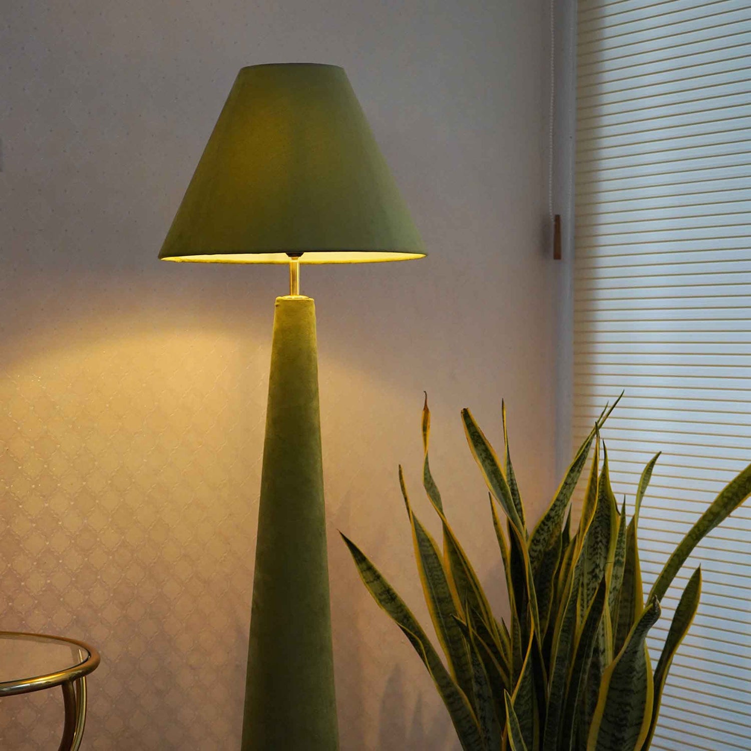 Green colored velvet floor lamp is lit-up and placed in a set-up with a large plant next to window blinds.