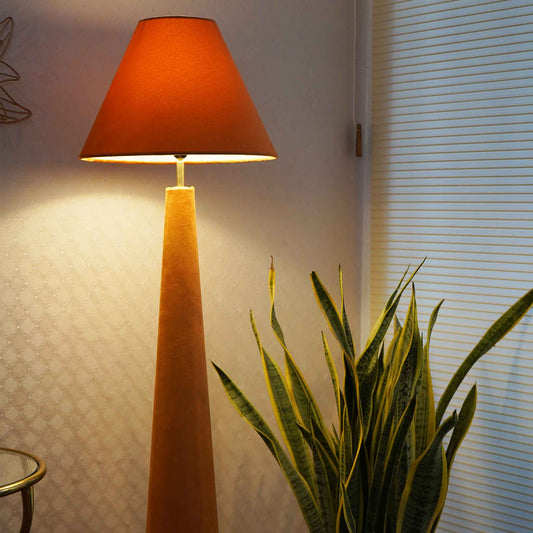 Orange colored velvet floor lamp is lit-up and placed in a set-up with a large plant next to window blinds.