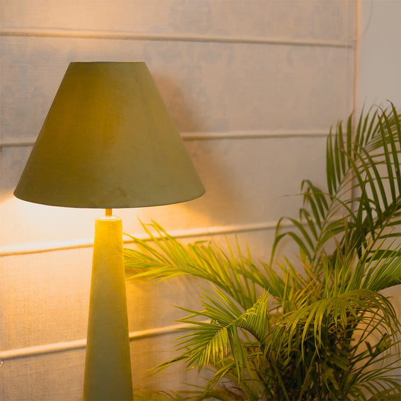 Green colored velvet floor lamp is lit-up and placed in front of a large indoor plant.